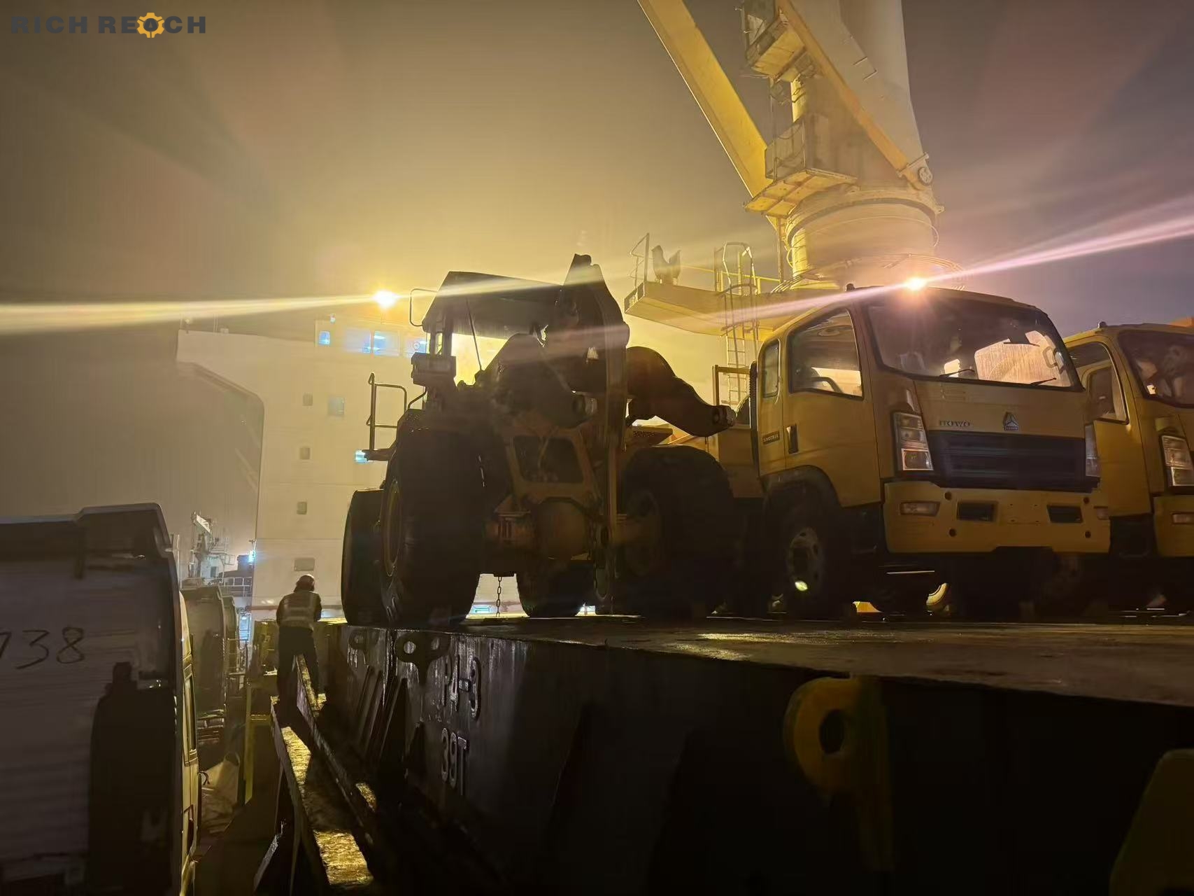 An excavator and yellow HOWO cargo trucks loaded on the deck of a freight ship at a port during a foggy night, with a harbor crane overhead.