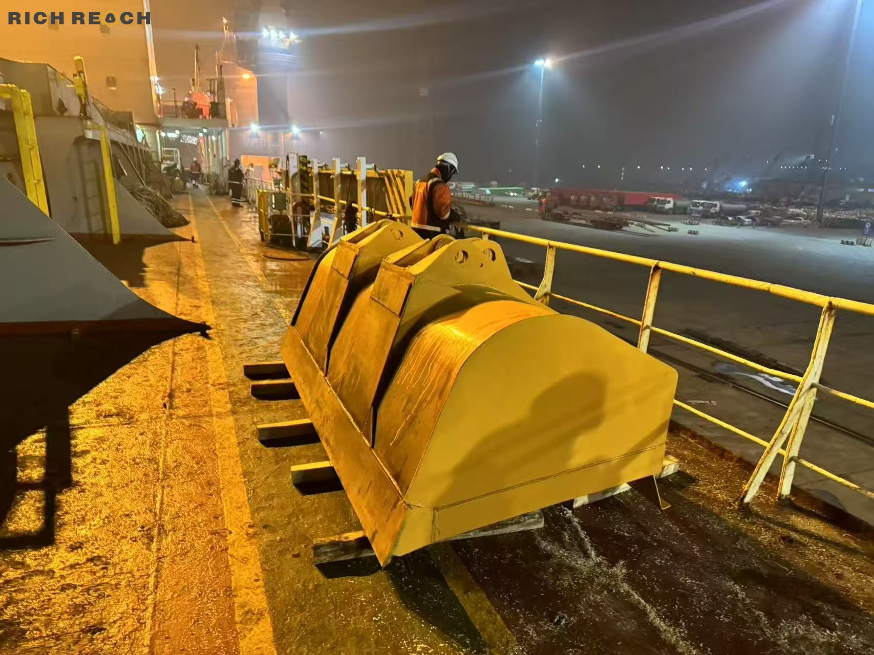 A large yellow heavy-duty excavator bucket secured on wooden blocks on the deck of a cargo ship at a port during a misty night.