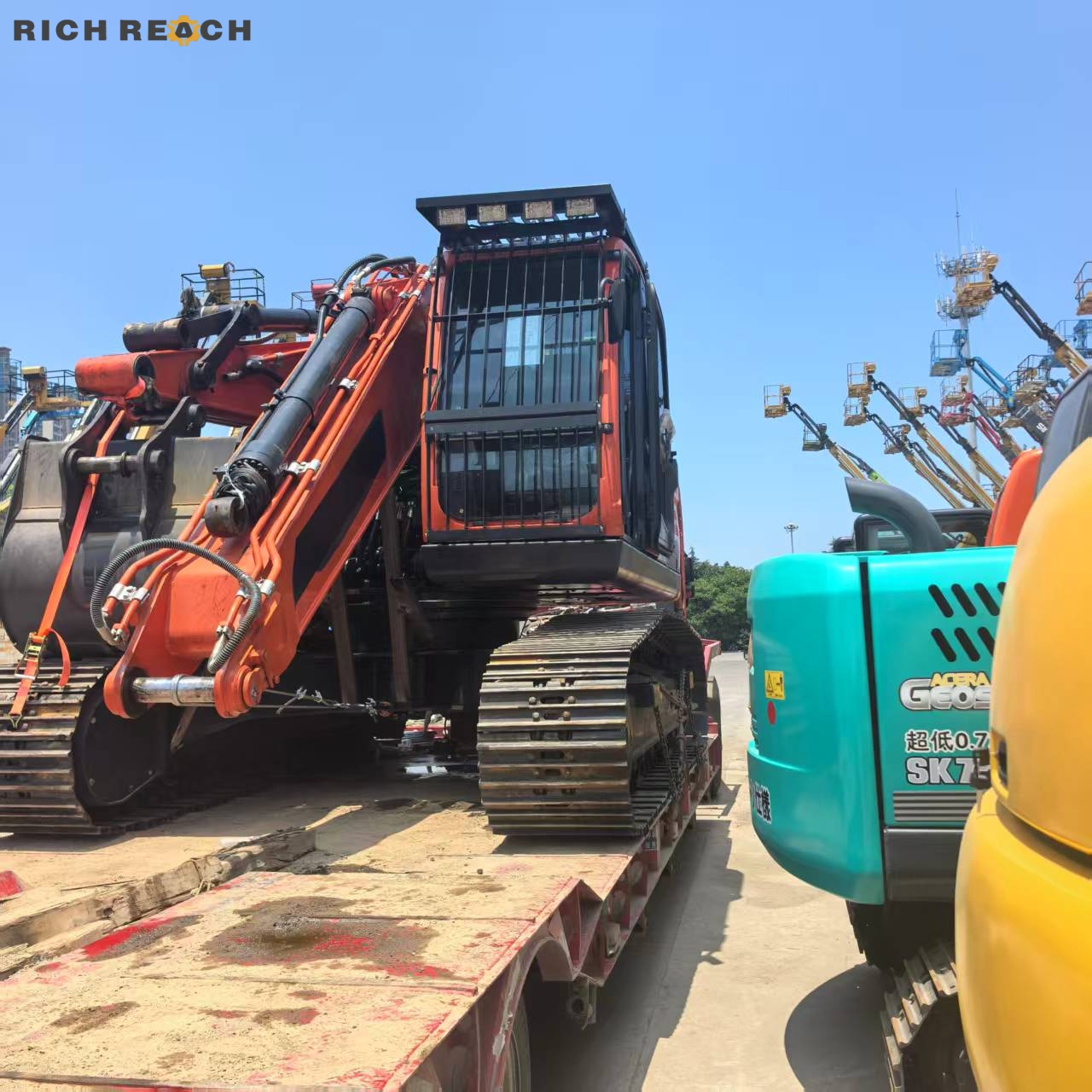 An orange crawler excavator with a protective cabin grid loaded on a flatbed semi-trailer at a machinery yard under a blue sky.