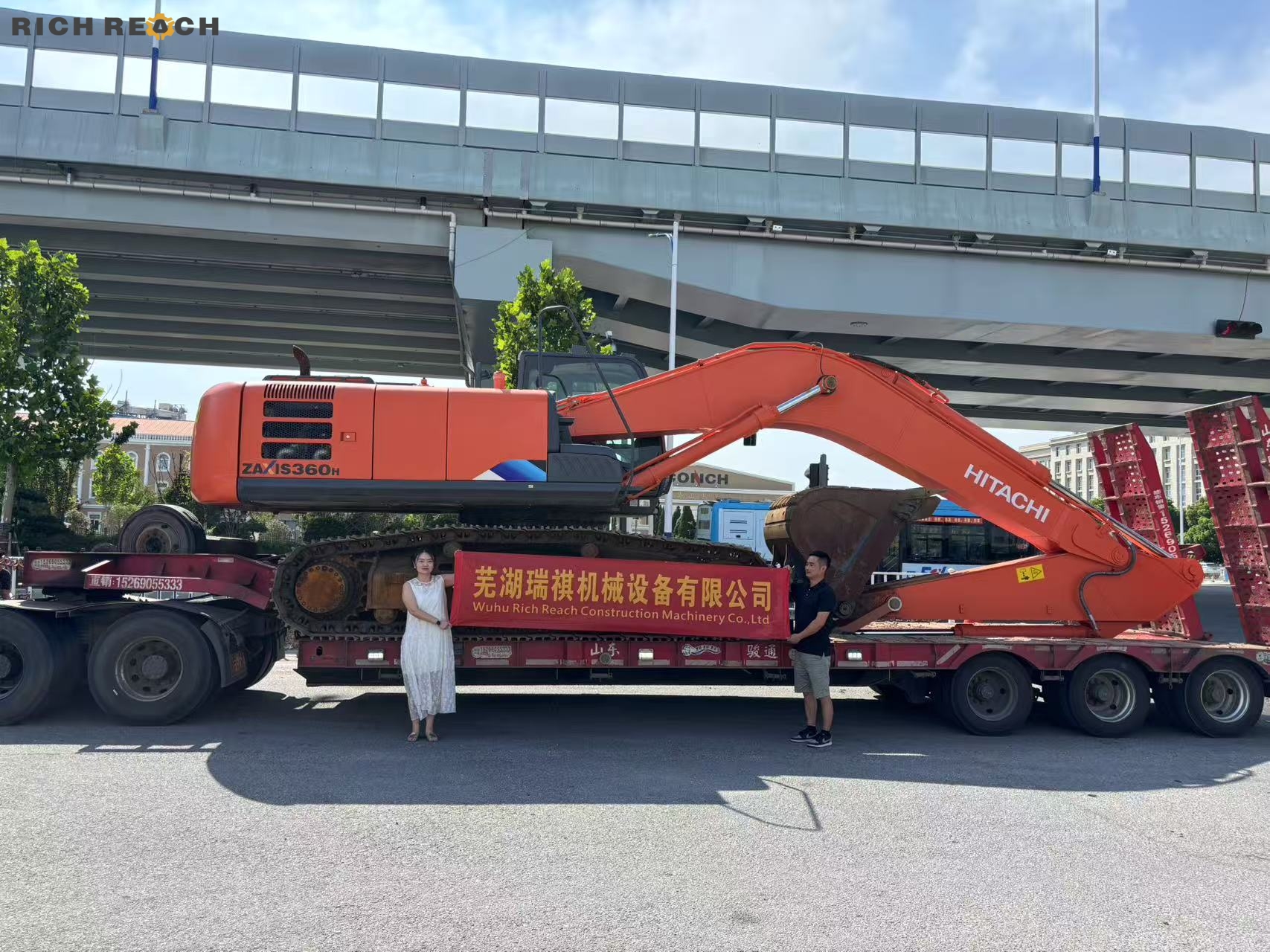 A large orange Hitachi ZAXIS 360H excavator loaded onto a heavy-duty flatbed semi-trailer, with two people holding a banner for Wuhu Rich Reach Construction Machinery Co., Ltd.