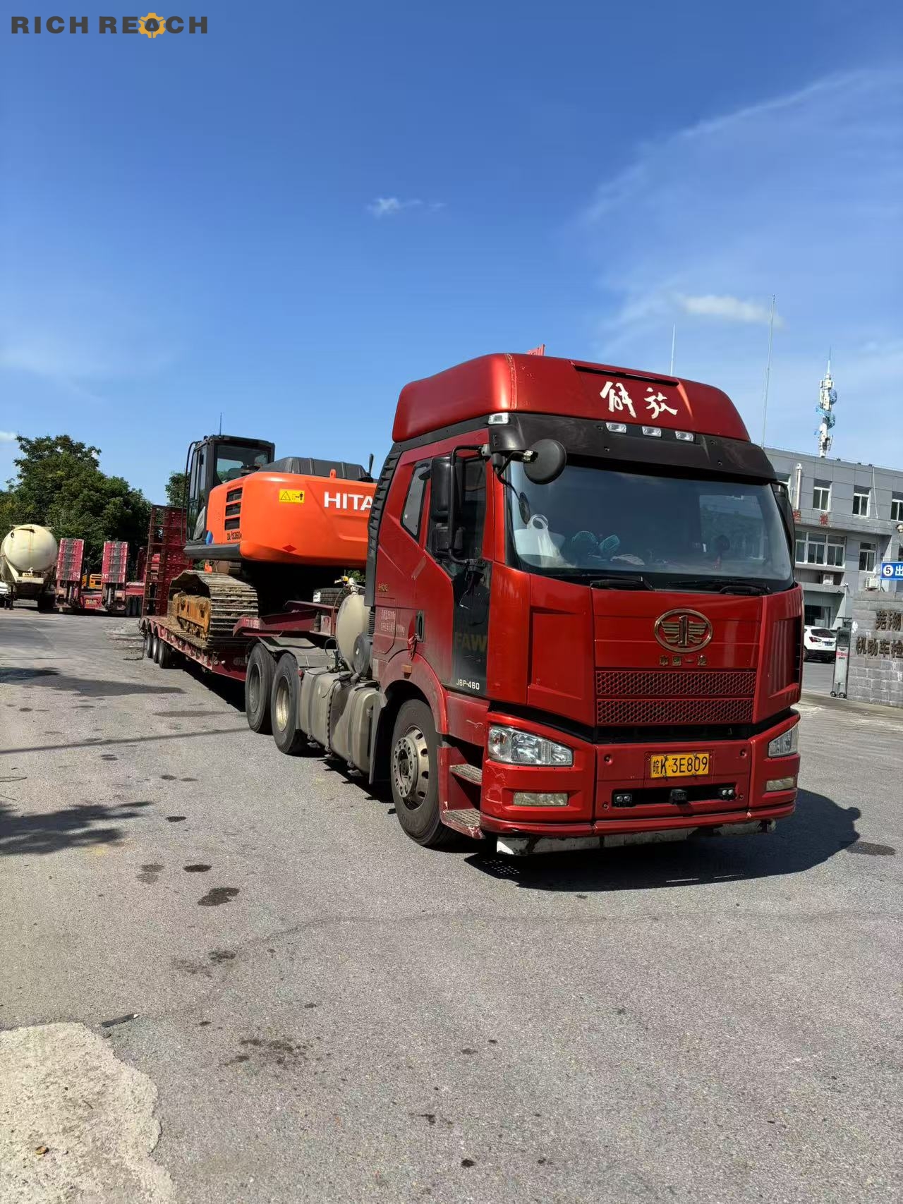 A red FAW Jiefang semi-truck pulling a lowbed flatbed trailer loaded with an orange Hitachi crawler excavator on an open road.