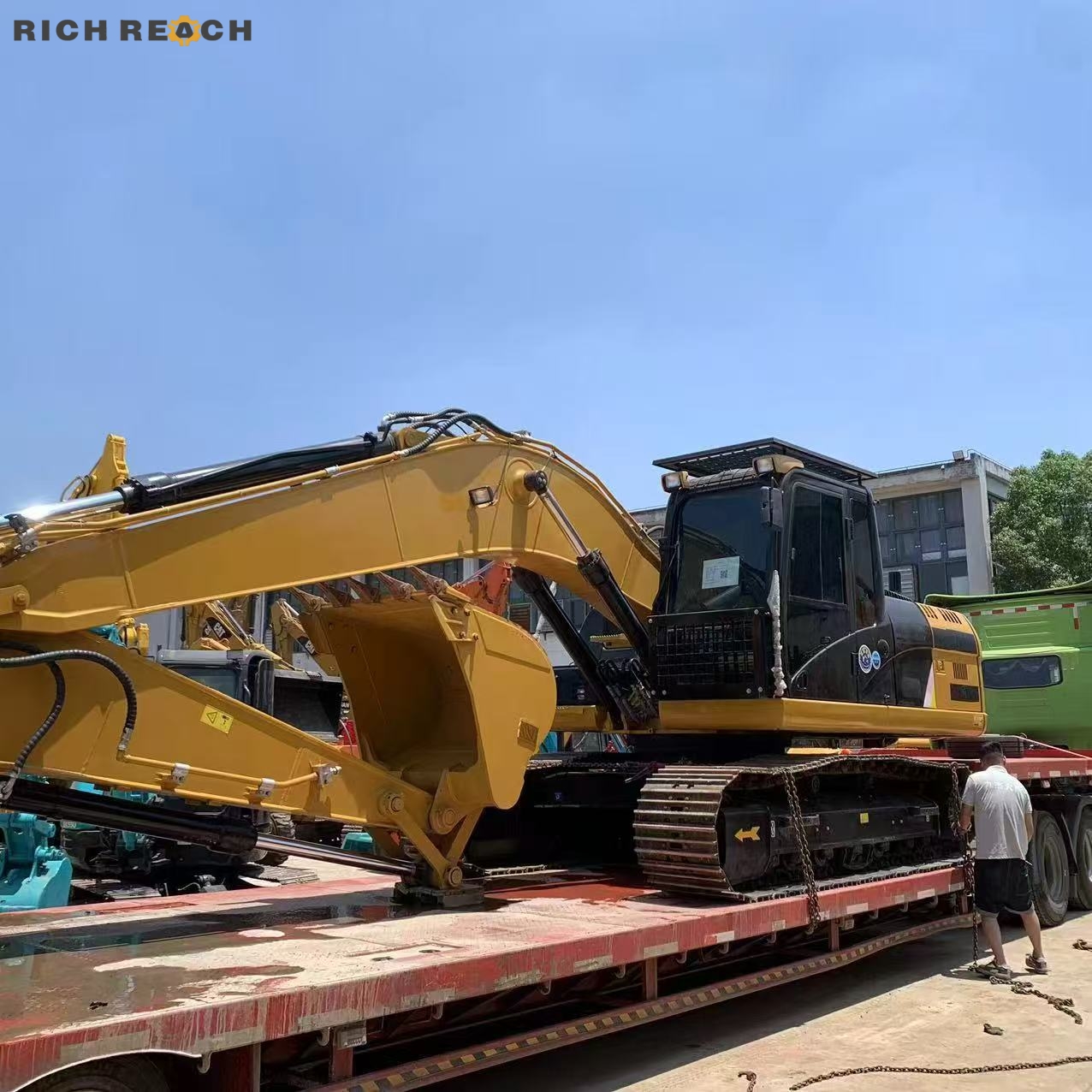 A yellow and black crawler excavator secured with heavy chains on a flatbed semi-trailer, with a worker checking the transport lashings under a clear sky.
