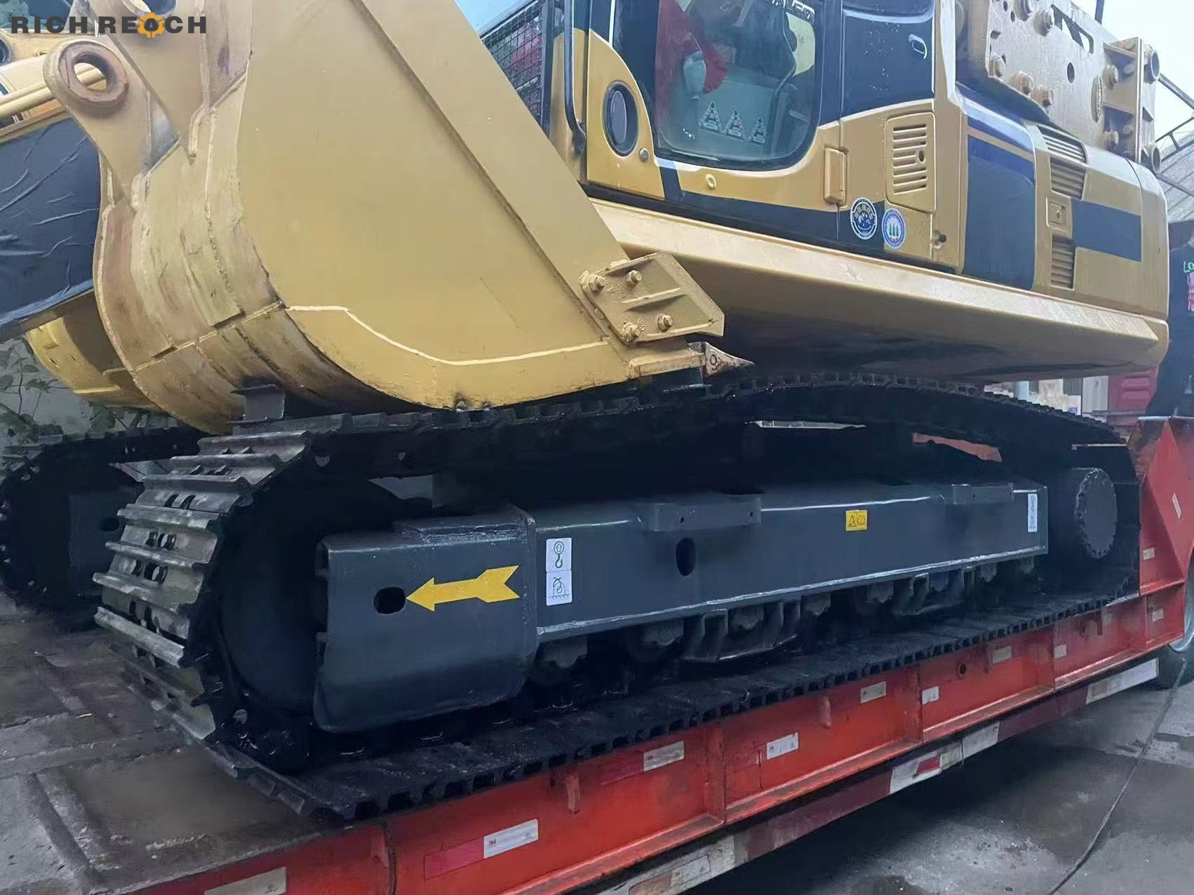 A close-up view of a heavy yellow crawler excavator's metal tracks and undercarriage secured on a red flatbed transport trailer.