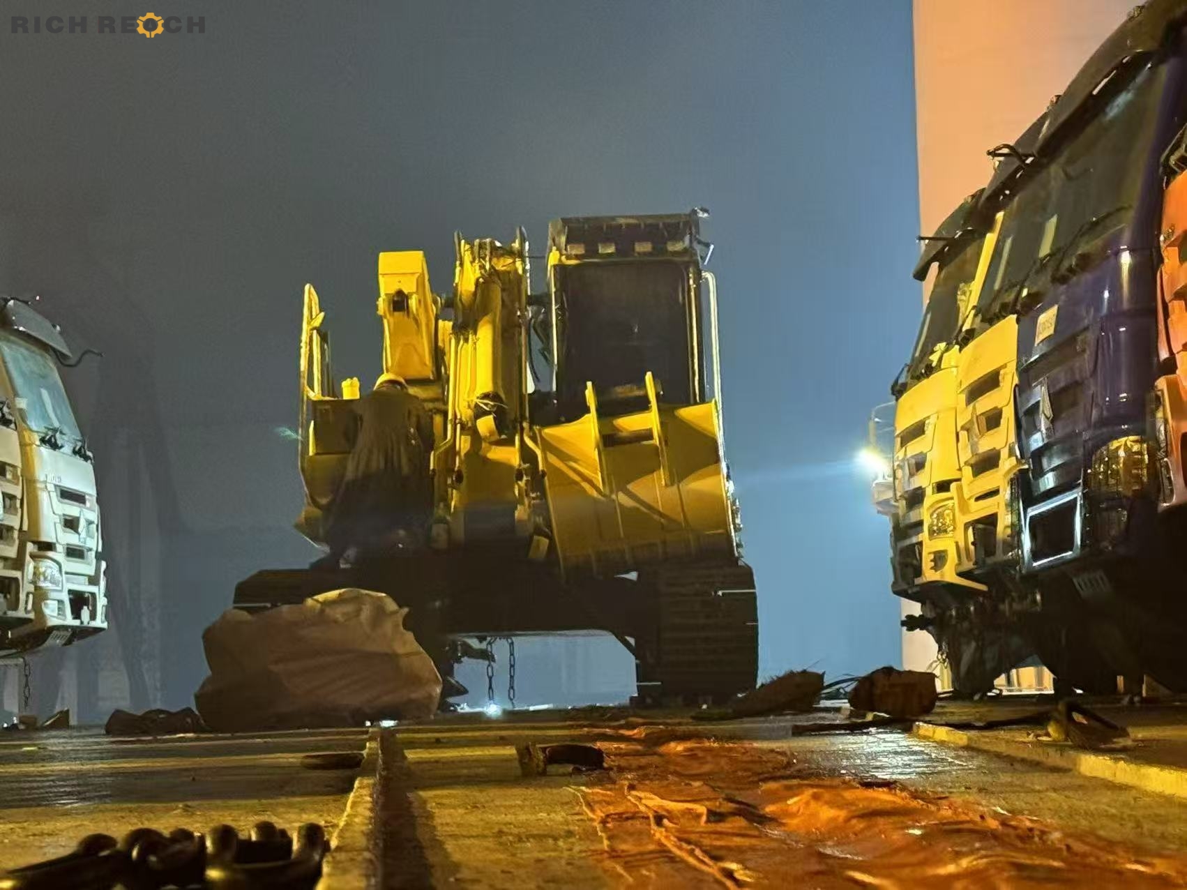 A yellow crawler excavator parked between commercial cargo trucks on the deck of a freight vessel or port terminal during a foggy night.